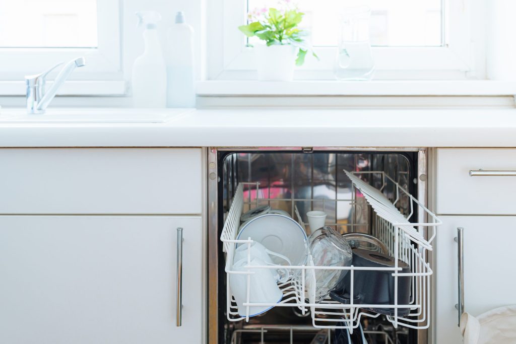 A built-in dishwasher under a window with the door open and the lower rack half out, plates and bowls standing in white racks