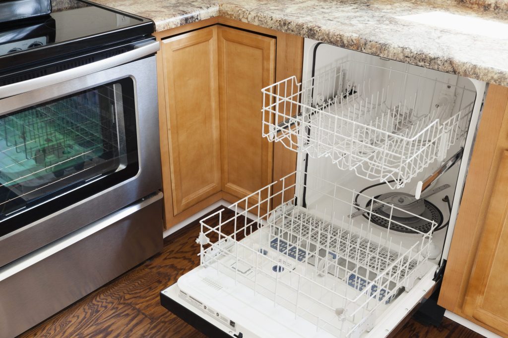 A built-in dishwasher with its door flat open and both white racks pulled out, oak cabinetry and a granite counter above, a range alongside