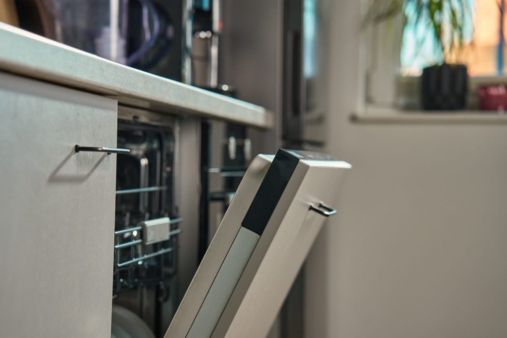 A built-in dishwasher with its door open a hand's width in a dark kitchen, the tub edge and the top of the racks showing
