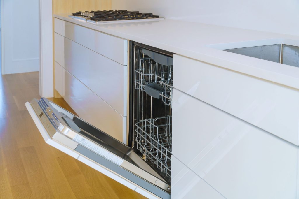 A built-in dishwasher plainly installed under a run of white cabinetry, seen at counter height with its door lowered and both racks in view