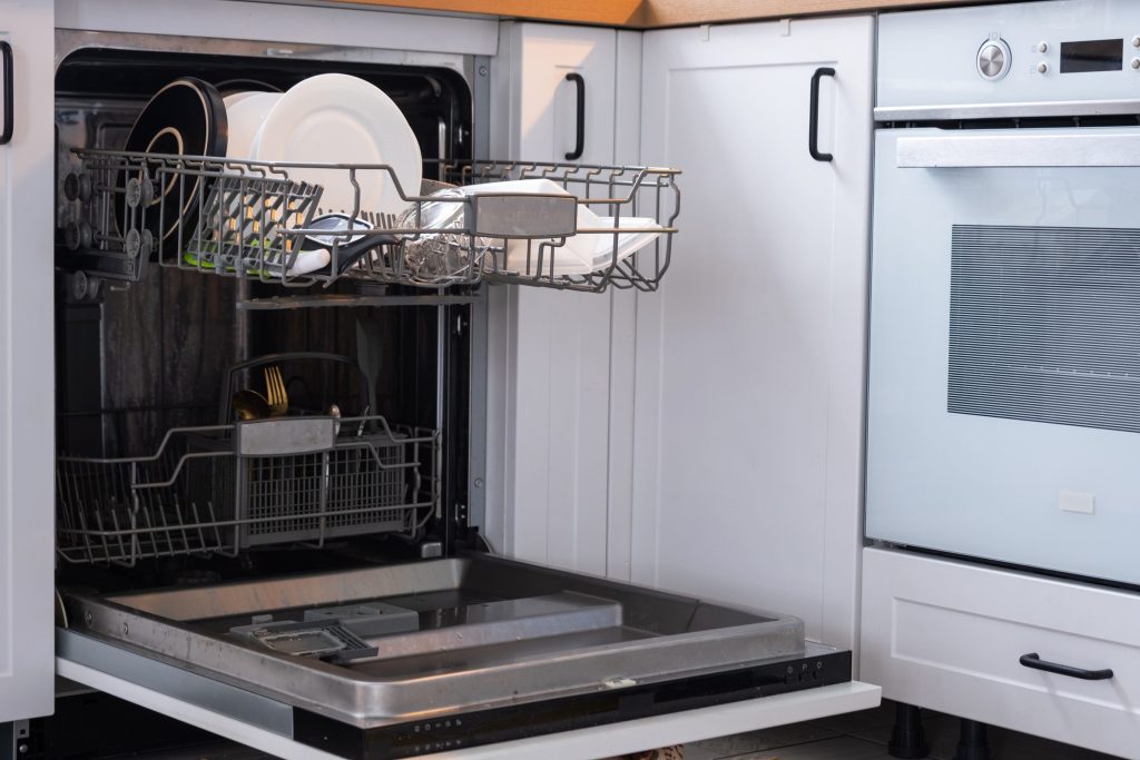 A built-in dishwasher with the door down and the upper rack pulled out, plates and bowls standing in it, a white wall oven alongside