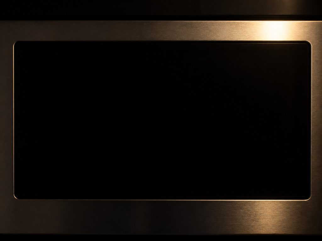 A close view of the digital display window on an appliance fascia in a dark kitchen, the screen completely dark and blank with no characters, digits or symbols of any kind on it, brushed metal around it holding one warm reflection, everything else in deep shadow.